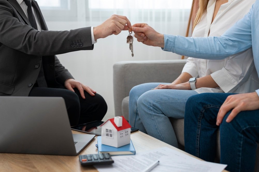 Deux personnes échangeant des clés de maison sur un bureau avec un agent immobilier et une maquette de maison sur la table.