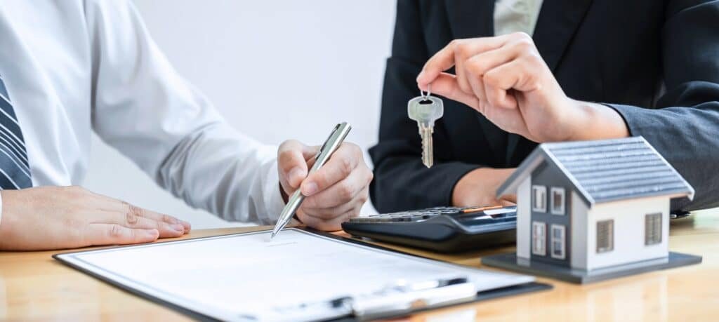 Agent immobilier remettant les clés d'une maison à un client lors d'une transaction immobilière, avec une maquette de maison et un contrat sur la table.