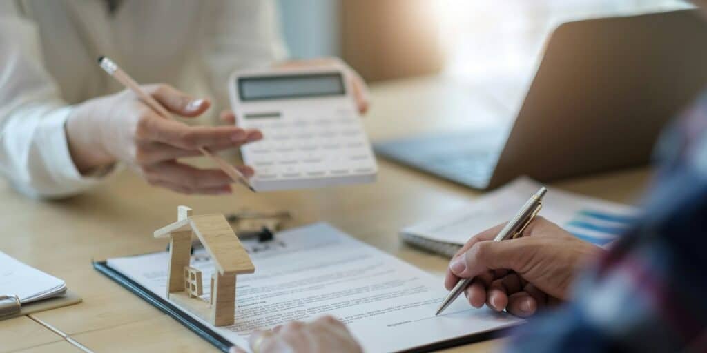 Deux professionnels discutant de calculs financiers avec un modèle de maison en bois sur la table.